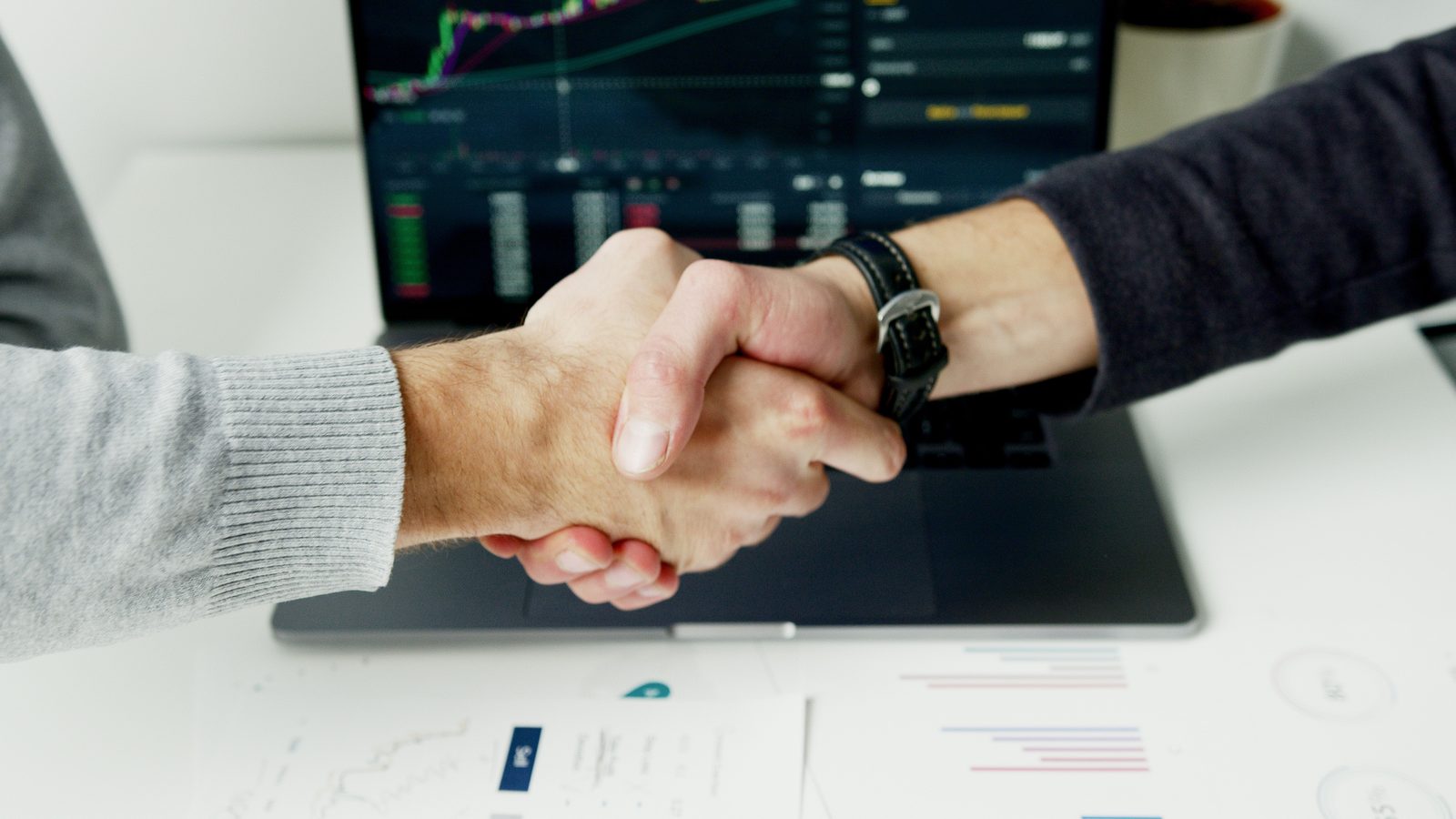 Two business professionals shaking hands over a desk with financial charts and reports, representing public-private partnerships